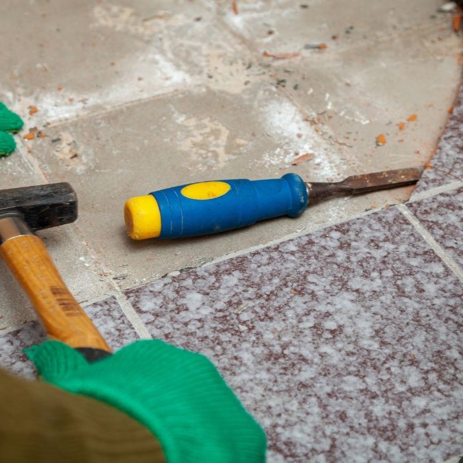 A chisel being hit with a hammer to loosen a tile attached to concrete.